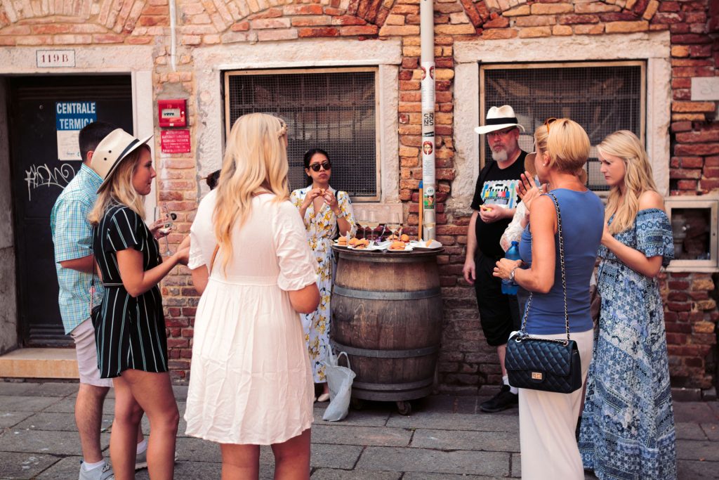 Several people in summer clothes stand around a barrel table with drinks and snacks, chatting on a cobblestone street by a brick building.