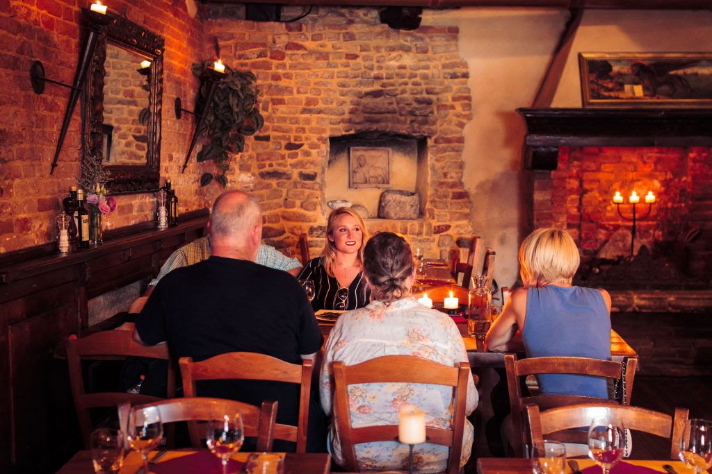 Five friends share a candlelit dinner at a rustic, brick-walled restaurant. One woman smiles warmly, facing the viewer.