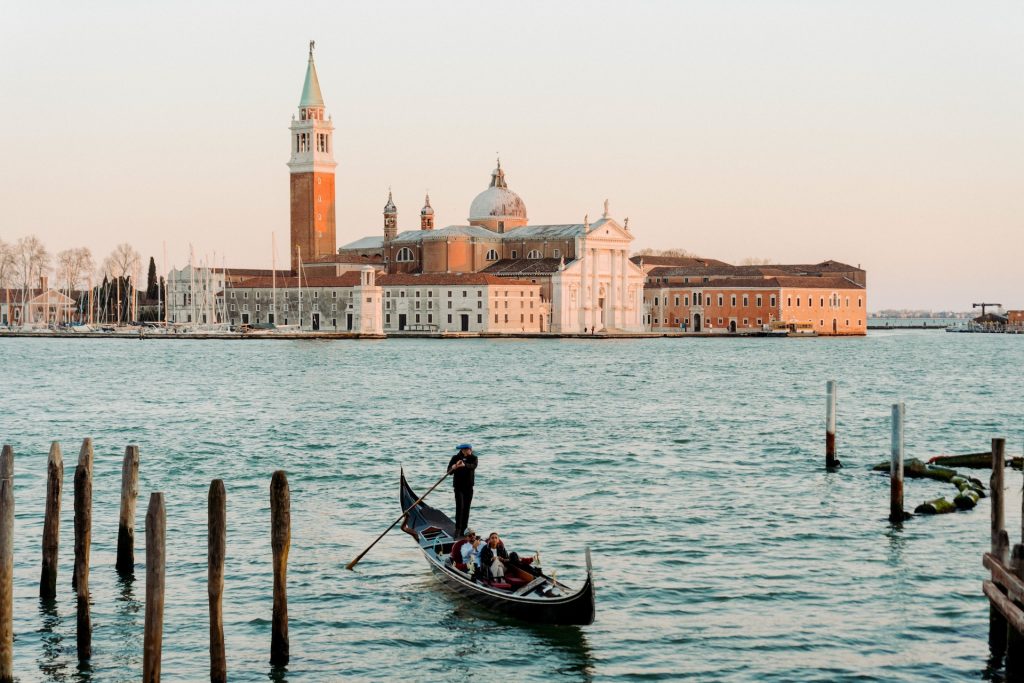 A gondolier rows a gondola with passengers across the water in Venice, passing San Giorgio Maggiore church and bell tower at sunset.