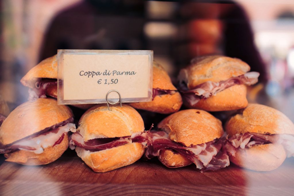 Crusty rolls filled with thinly sliced Coppa di Parma are stacked in a display case in Italy, priced at €1.50 each.