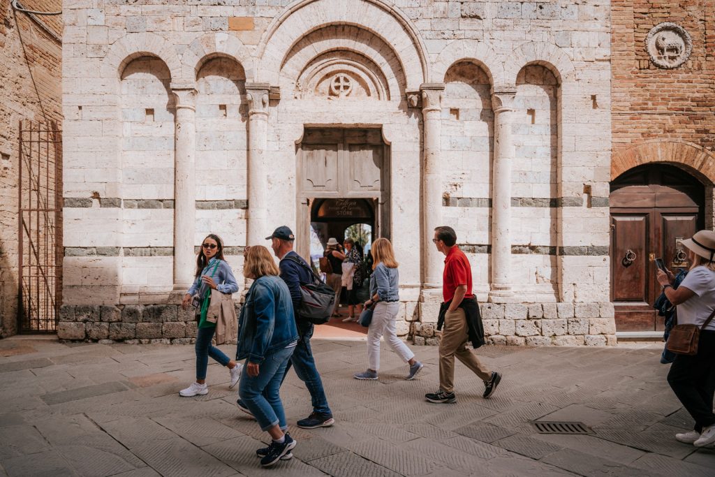 Tourists stroll a bustling cobblestone street in Tuscany, pausing by a historic stone church with striped arches and ornate doorways.