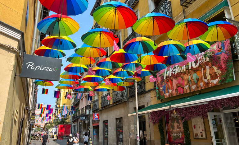Colorful rainbow umbrellas and pride flags hang above a lively street in Chueca, the Madrid gay district, with LGBTQ+ venues and shops visible below.