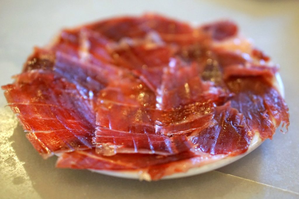 Thin slices of cured Jamón Ibérico are neatly layered on a white plate, served as traditional tapas in Seville, Spain.