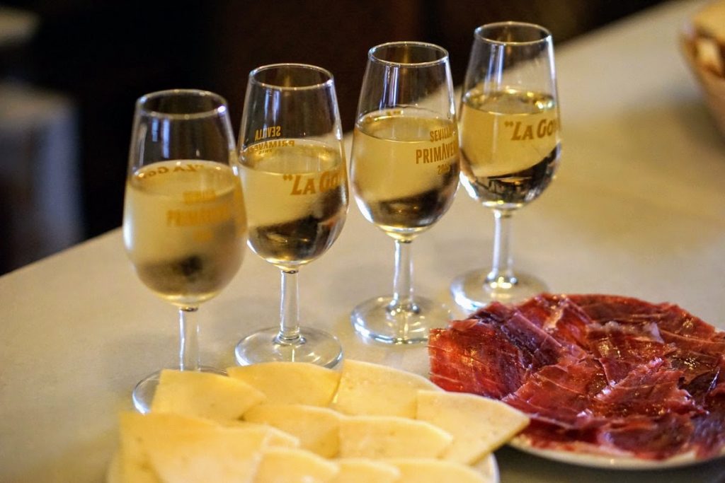 Four glasses of white wine sit on a wooden table in Seville, surrounded by plates of Jamón Ibérico and Manchego cheese.