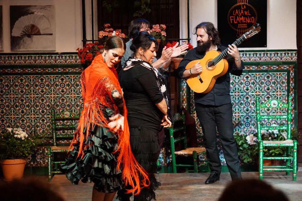 Two women in vibrant flamenco dresses dance on a Seville stage, accompanied by clapping and guitar in Casa el Flamenco.