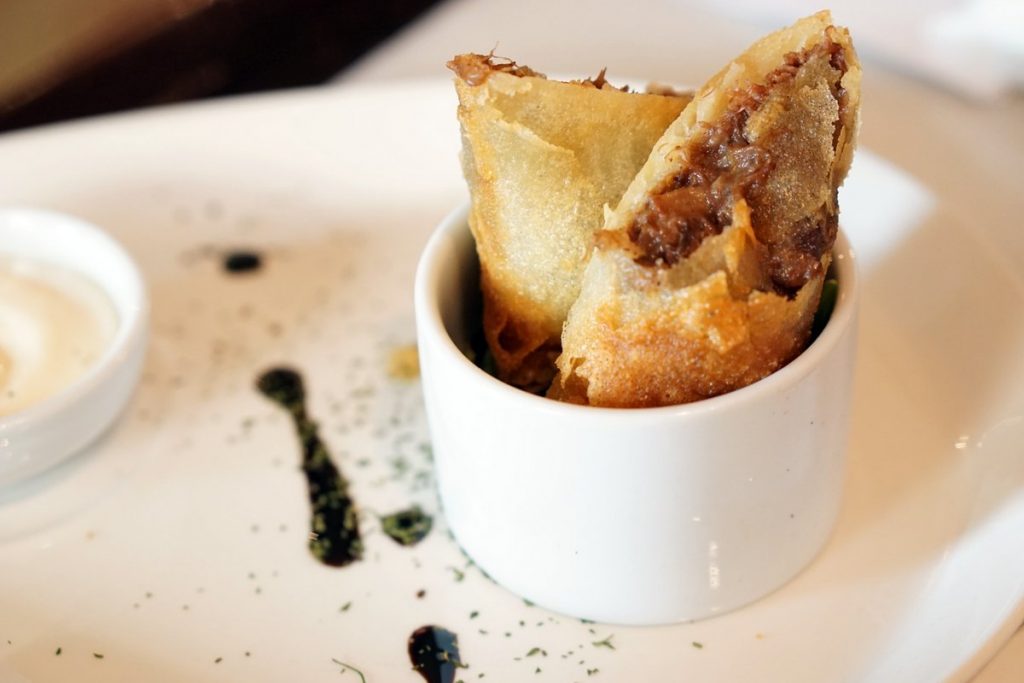 Two golden spring rolls stand upright in a white ramekin, garnished with fresh herbs and served alongside a dish of dipping sauce.