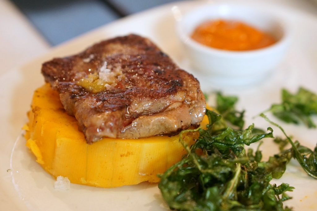 A seared steak rests on roasted squash, garnished with flaky salt, crispy greens, and orange sauce, served in Seville-style presentation.