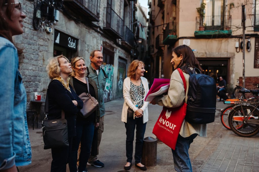 Five people smile on a narrow street while a woman with a backpack and tote bag gives restaurant recommendations; bicycles nearby.