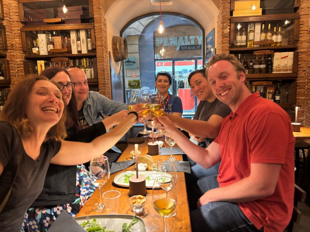 Six friends raise wine glasses in a Naples restaurant, enjoying dinner together. Wine bottles and an arched window set a cheerful scene.