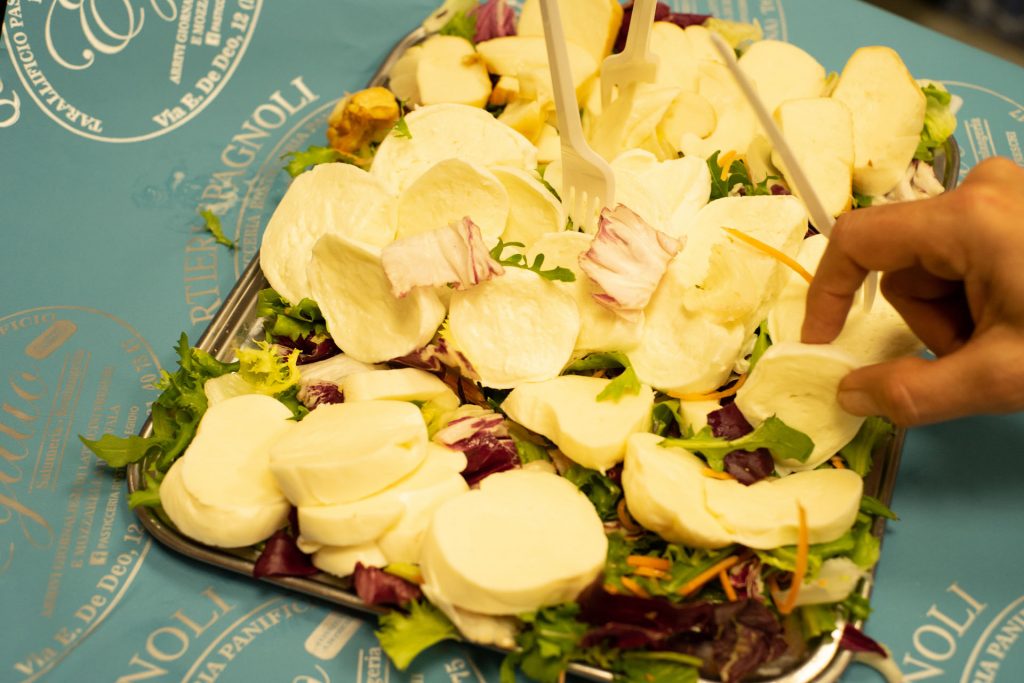 A hand reaches for fresh mozzarella slices atop mixed greens, with plastic forks, on a blue tablecloth in Naples, Italy.