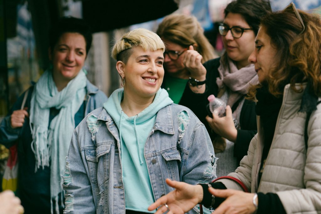 Several women gather outdoors in Naples, chatting and smiling. The woman in front wears a teal hoodie and ripped denim jacket.