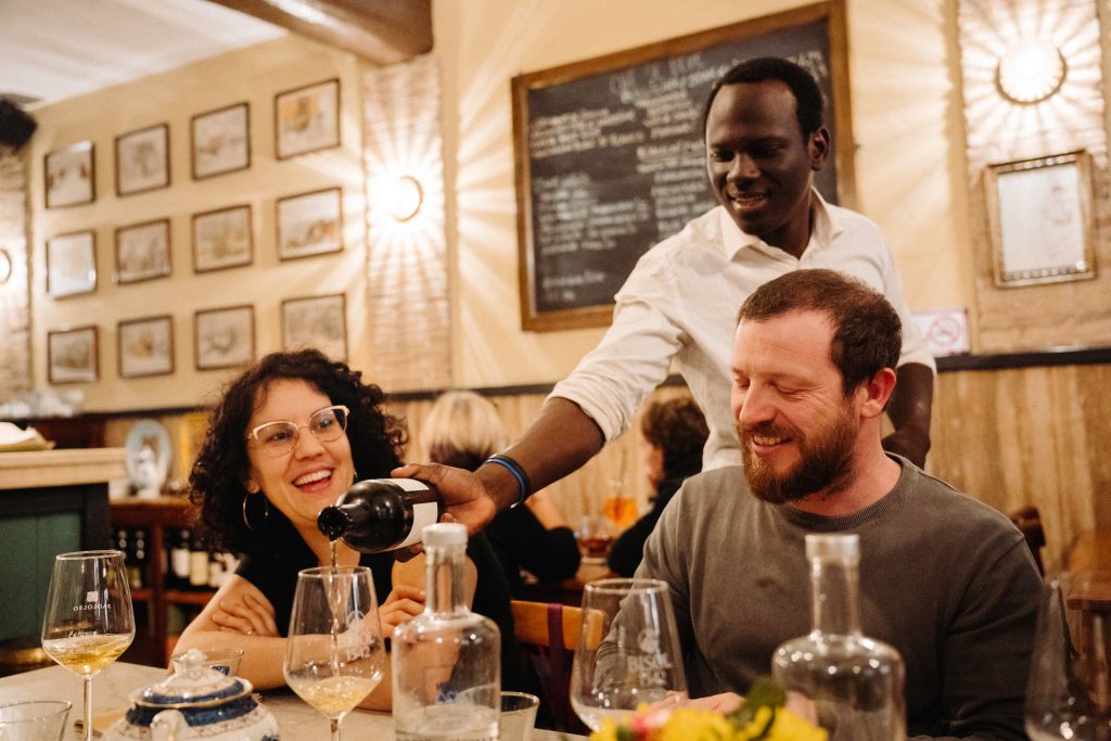 A waiter pours wine for a couple dining at a warmly lit Naples restaurant, with framed artwork and several wine bottles nearby.