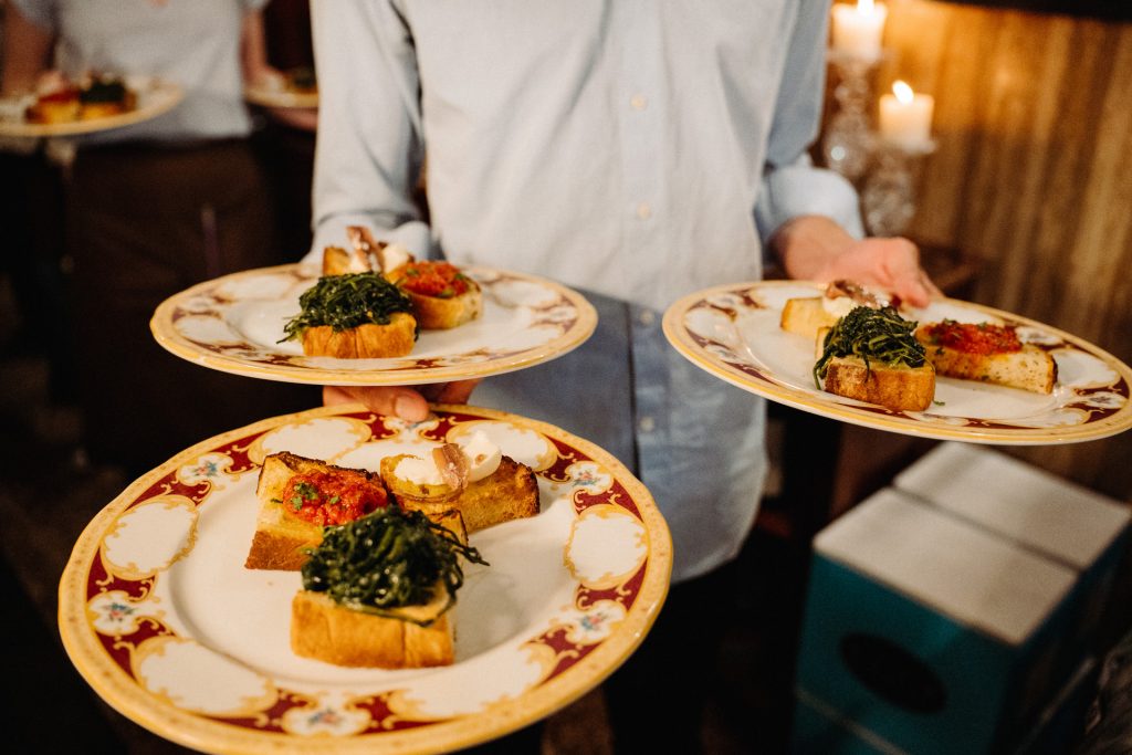 A server in a light blue shirt offers three plates of bruschetta appetizers at an indoor Naples dining event with warm lighting.