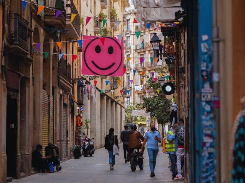 A lively street in Barcelona’s gay neighborhood features colorful banners, a large pink smiley sign, people walking and riding scooters.