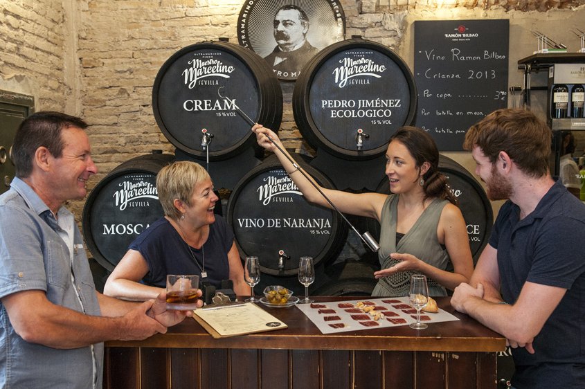 Four friends laugh and sip wine in a rustic cellar in Seville, surrounded by labeled barrels and a chalkboard tapas menu.