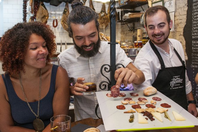 Three friends share a platter of cured meats, cheeses, and bread in a cozy Seville deli; one reaches for salami.