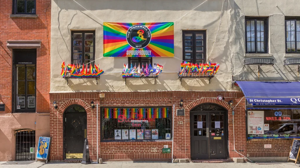 The Stonewall Inn in New York displays rainbow Pride flags and a #ProudProudly banner, celebrating LGBTQ+ pride and history.