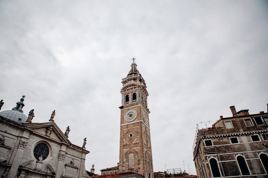 A tall clock tower with a cross rises among ornate historic buildings in Venice, featuring statues, carvings, and a large round clock.