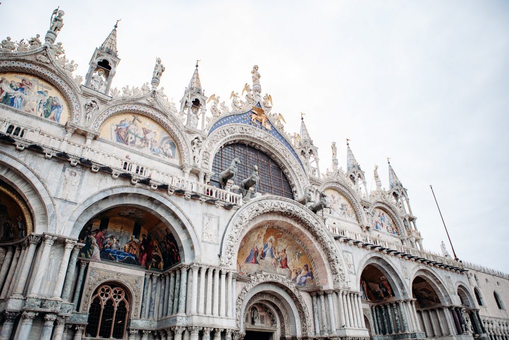 St. Mark’s Basilica in Venice, Italy displays arched doorways, detailed mosaics, columns, and statues below a cloudy sky.