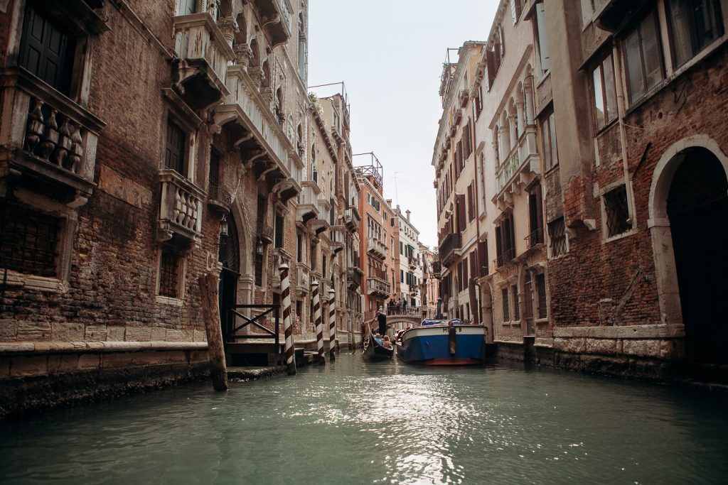 A narrow canal in Venice, Italy features a blue boat and gondola floating between historic, weathered buildings under soft sunlight.