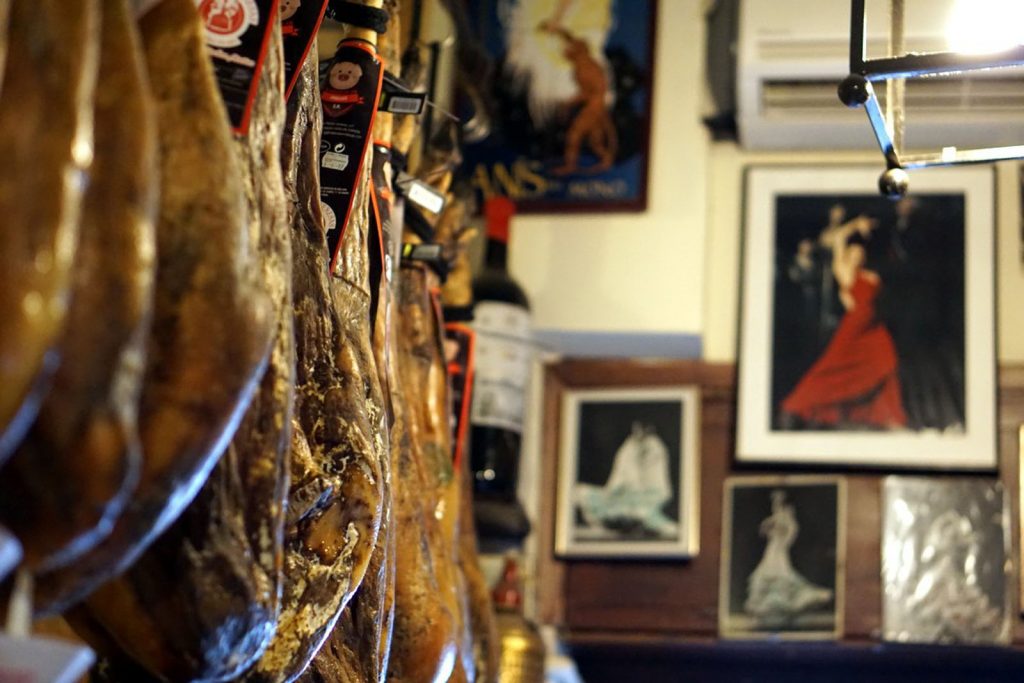 Several cured meats hang from the ceiling of a Seville bar, with flamenco dancer portraits and wine bottles in the background.