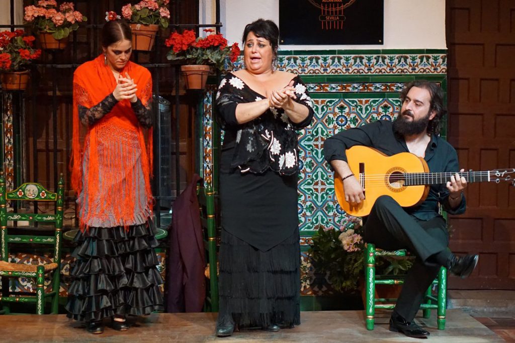 Three flamenco performers in Seville: a woman claps, another sings passionately, and a bearded man plays guitar before colorful tilework.