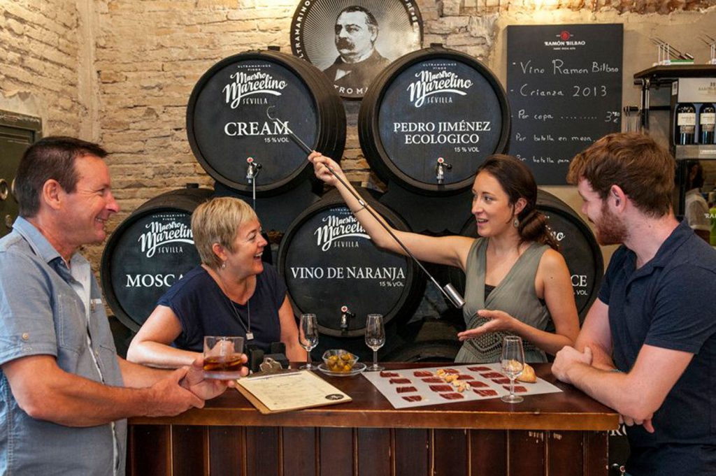 Four friends share drinks and laughter in a rustic Seville wine bar, as a woman enthusiastically pours red wine from a barrel.