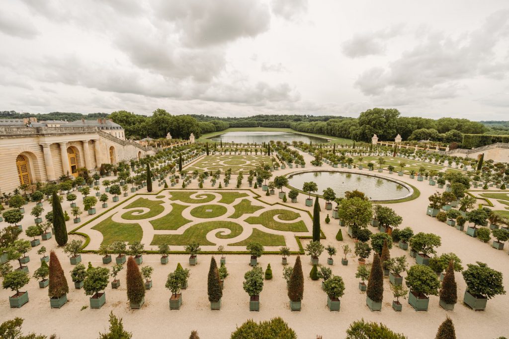 An ornate formal garden at Versailles features intricate hedges, circular ponds, potted trees, and the Palace under cloudy skies.