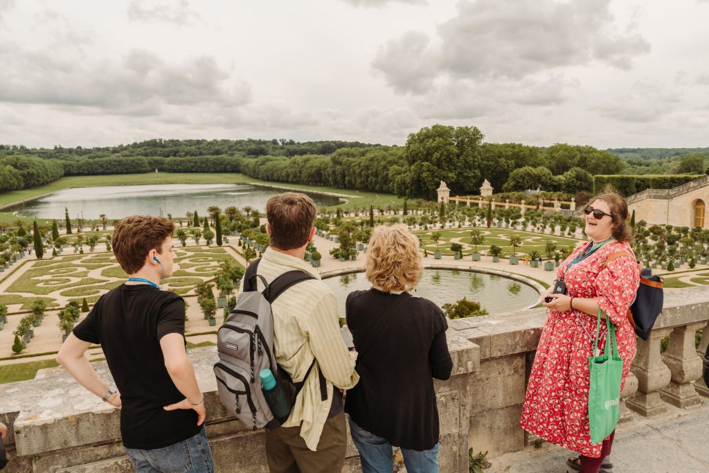 Four people stand on a stone terrace at Versailles, listening to a woman in a red dress explain the grand formal gardens below.