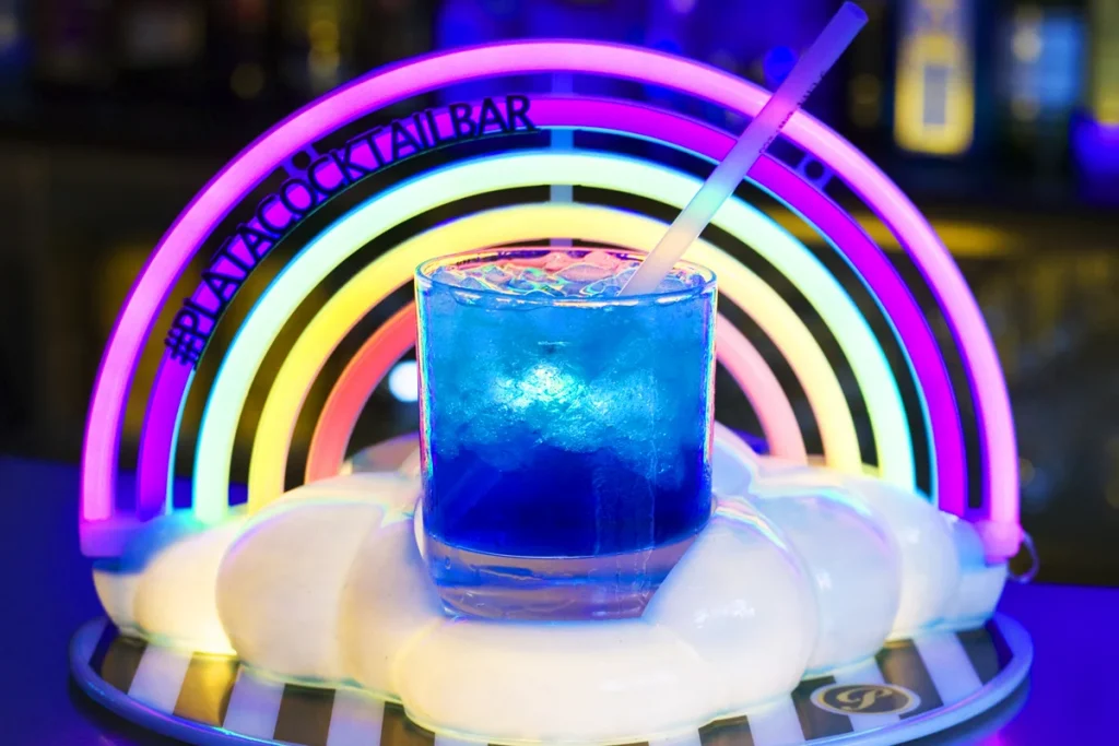 A blue cocktail with ice sits on a cloud tray beneath a neon rainbow at #PLAYACOCKTAILBAR in Barcelona’s gay district.