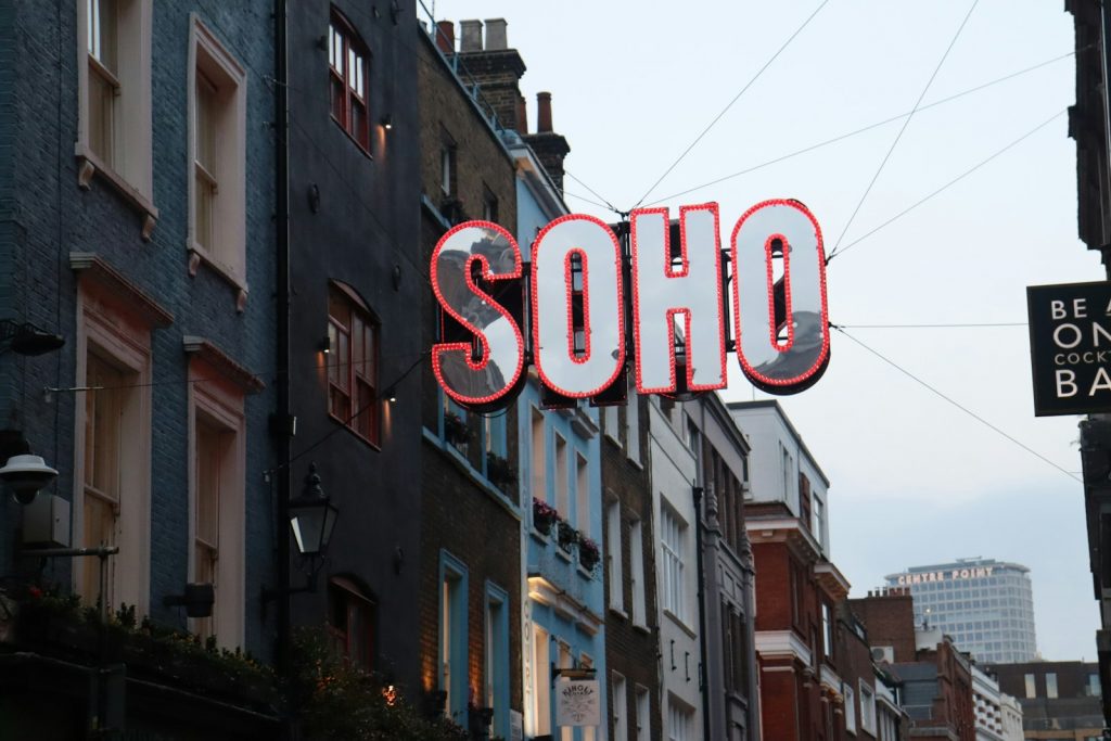 A large illuminated "SOHO" sign hangs above a lively London street, surrounded by colorful buildings and city atmosphere.