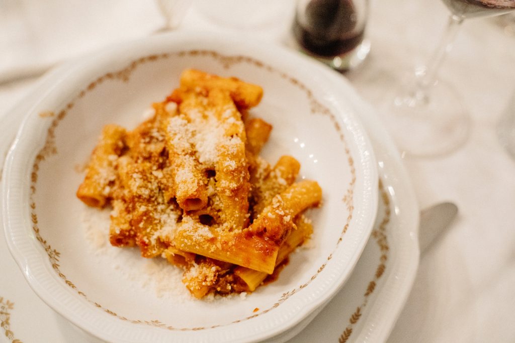 A white ceramic bowl holds rigatoni pasta in tomato sauce with grated cheese. A glass of red wine sits behind it.