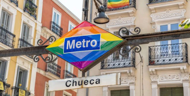 Rainbow-themed Chueca Metro sign in Madrid’s gay district, with traditional Spanish architecture and LGBTQ+ pride flags in the background.