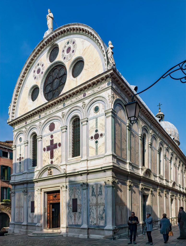 The marble facade of Santa Maria Novella in Florence features statues, arched windows, and visitors gathered under a bright blue sky.