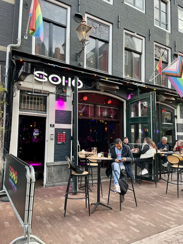People relax at outdoor tables under rainbow flags at Soho café in Amsterdam’s Gay District, enjoying a lively, LGBTQ+ inclusive atmosphere.