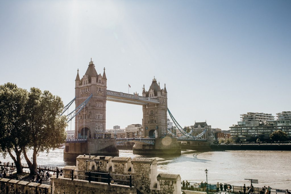 Tower Bridge stretches over the River Thames in London; pedestrians stroll nearby, with modern skyscrapers and bright sunlight illuminating the scene.