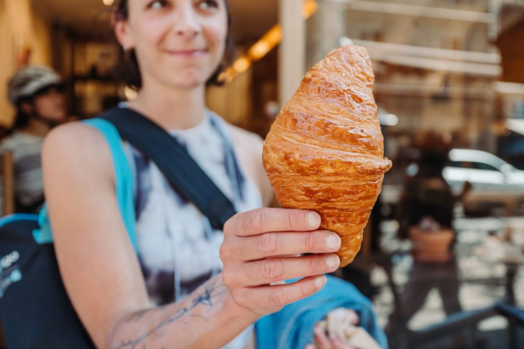 A person smiles, presenting a large, flaky croissant in a Parisian café, celebrating the delight of French pastry.
