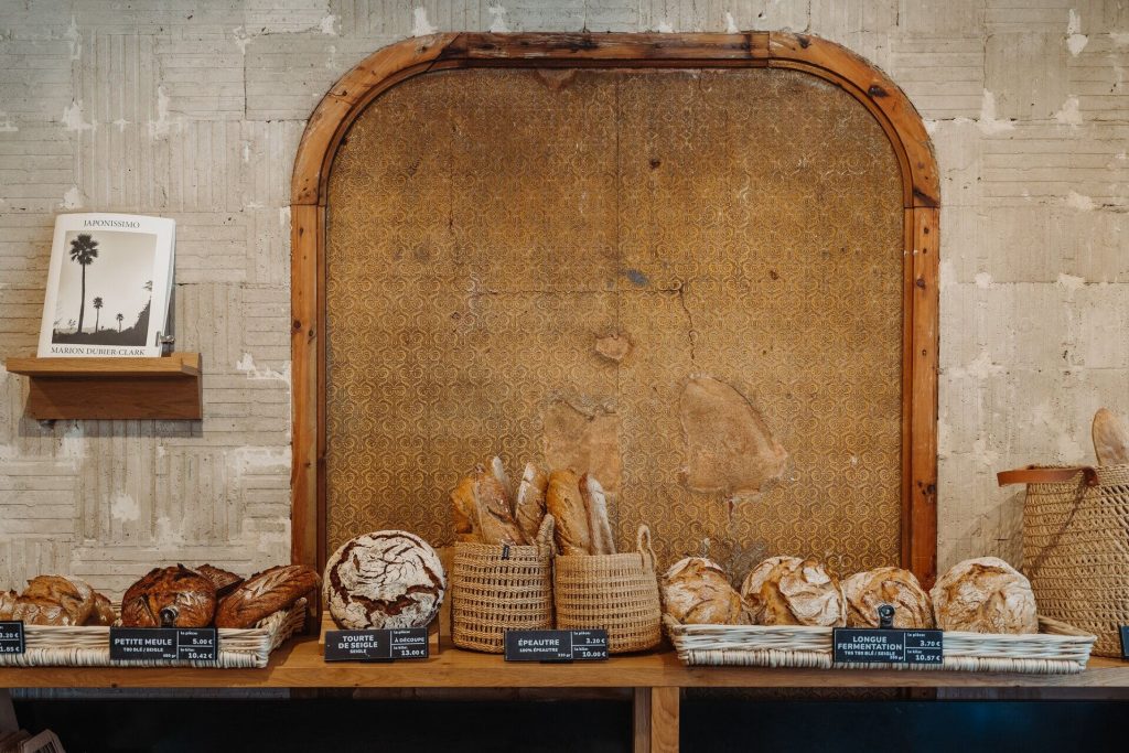 A wooden shelf inside a Parisian bakery displays assorted rustic breads in baskets and trays, each labeled; a cookbook rests above.