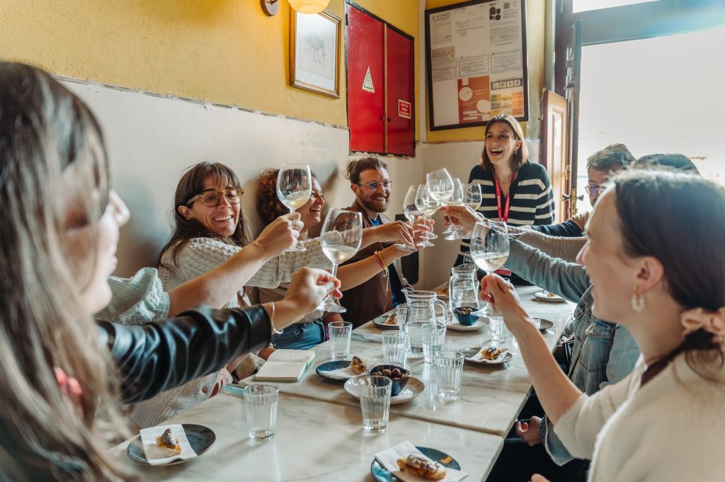 Friends gather at a sunny restaurant table, laughing and toasting with drinks. Plates of food and water fill the table.