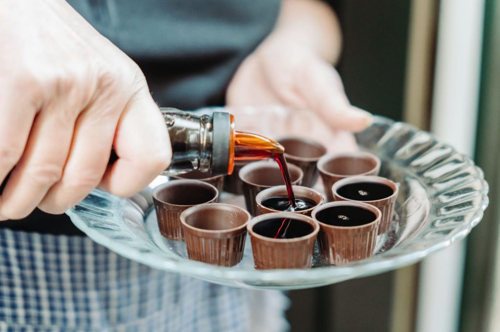 A hand pours dark liqueur from a bottle into several chocolate cups neatly arranged on a glass platter.