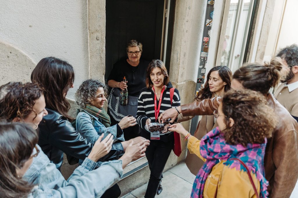 Several people stand outdoors in a circle, reaching for chocolates from a smiling woman with a red lanyard. A woman observes nearby.