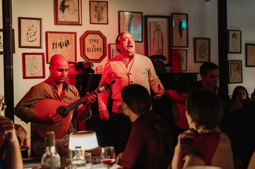 A man sings on stage as two musicians play guitars. Diners watch in a warmly lit restaurant decorated with framed paintings.