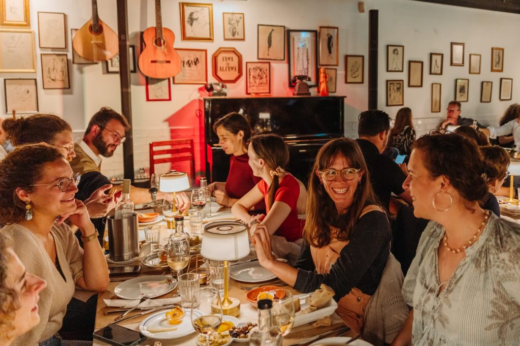 Several friends share food and drinks at a cozy restaurant with framed paintings and guitars on the warmly decorated walls.