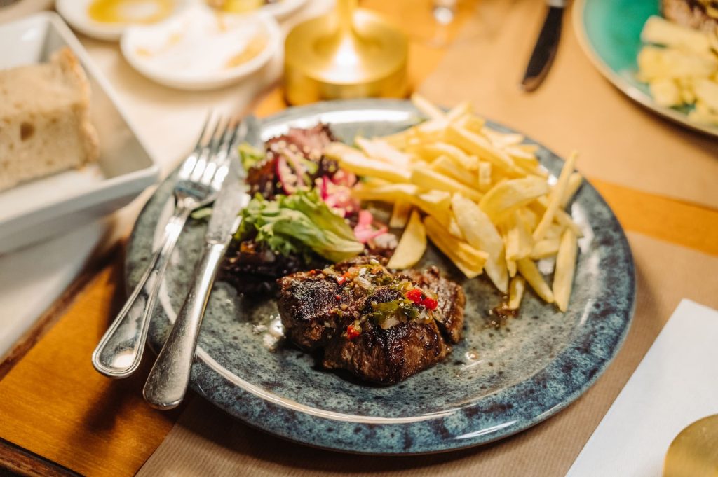 Grilled steak with herbs, French fries, and salad sit on a plate. Fork, knife, bread, and other dishes surround the meal.
