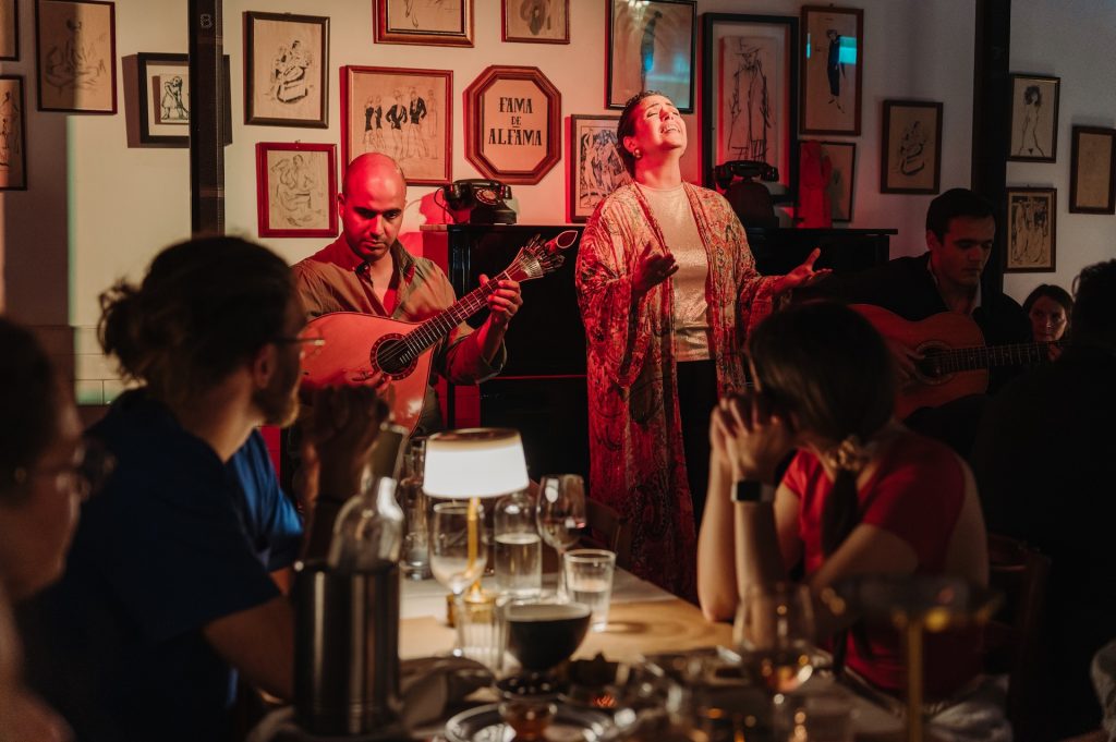 A singer performs with guitarists for an attentive group in a warmly lit restaurant decorated with framed art on the walls.