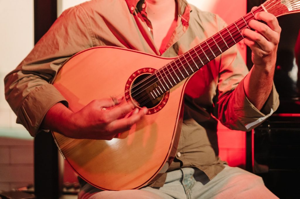 A person in a button-up shirt expertly plays a Portuguese guitar under warm red lights on stage, focused and expressive.
