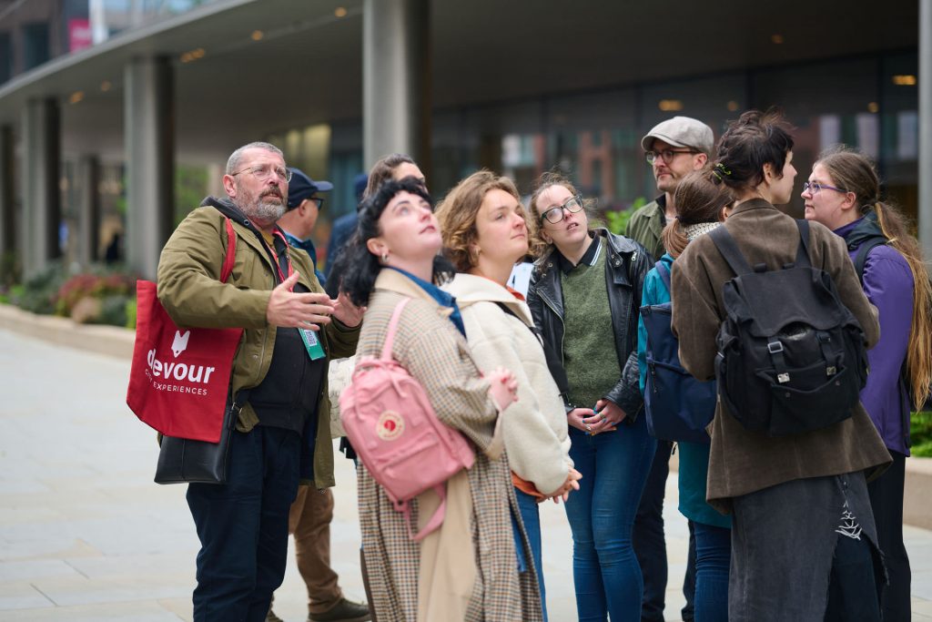 A tour guide with a red tote bag speaks to an attentive group during a London pub crawl, as participants listen closely.
