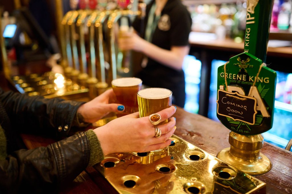 A person holds two pints of beer at a busy London pub, with brass taps and a Greene King IPA tap.