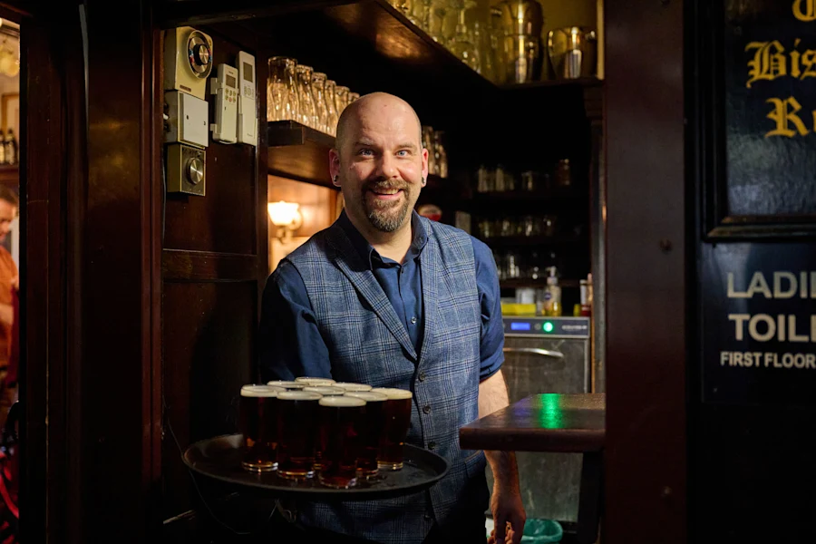 A smiling bartender in a blue vest serves beer from behind the bar at a warmly lit London pub, shelves behind him.