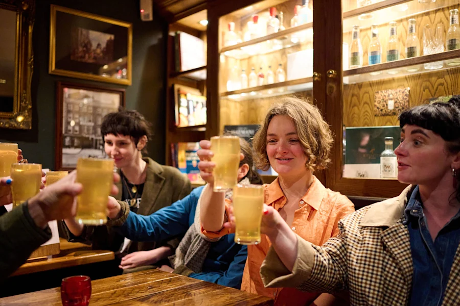 Several friends sit at a wooden table in a cozy London pub, smiling and toasting with glasses of beer and cider.